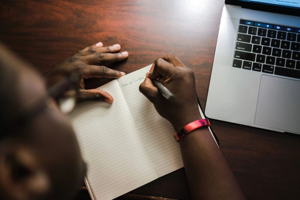 A person writing on paper with a laptop in the background.