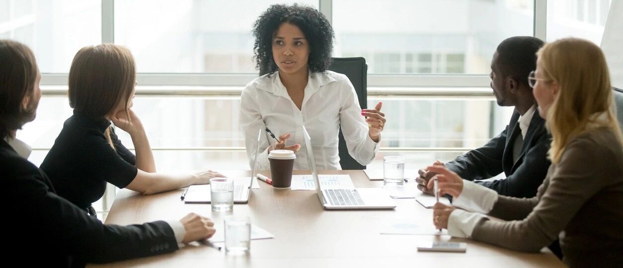 A woman in a white shirt speaking during a meeting with colleagues.